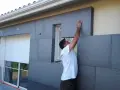 Un ouvrier pose des panneaux isolants gris pour l'isolation par l'extérieur d'une maison, près d'une fenêtre avec volet roulant.
