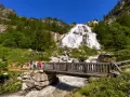 Cascata imponente con escursionisti su un sentiero, un ponte di legno attraversa un torrente. Un esempio di cosa vedere in Piemonte per gli amanti della natura.