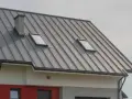 Nowoczesny dach z szarej blachy na rąbek, z dwoma oknami dachowymi i kominem.
