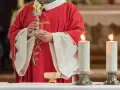 Kapłan w czerwonej szacie z krzyżem trzyma kielich podczas konsekracji. Płoną dwie świece.