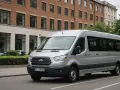 Un homme est au volant, prêt à conduire un minibus argenté dans une rue bordée d'arbres et de bâtiments.