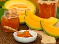Marmellata di melone in vasetti, con fette di melone fresco e un cucchiaino pieno di confettura.