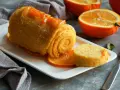 Pomarańczowy deser w rolce, posypany cukrem i ozdobiony skórką pomarańczy. Obok plasterki owocu i widelec.
