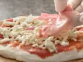 Ręka w białej rękawiczce kładzie plasterki szynki na pizzy z serem i sosem pomidorowym.