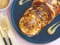 Goldbraune Quarkkeulchen ohne Kartoffeln, bestreut mit Zucker, auf einem dunklen Teller mit Apfelmus. Daneben liegen goldene Besteckteile.