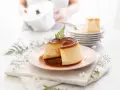 Delicioso flan de queso con caramelo líquido, decorado con una hoja verde. En el fondo, una persona prepara más postres.