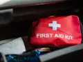 Czerwona apteczka samochodowa z białym krzyżem i napisem "FIRST AID KIT" leży w schowku. Obok niej papierowy ręcznik.