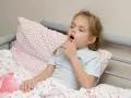 Mała dziewczynka z **przewlekłym kaszlem u dziecka** leży w łóżku, trzymając termometr.
