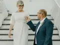 Elegancka kobieta po pięćdziesiątce w sukience do ślubu cywilnego, trzymająca za rękę pana młodego w granatowym garniturze.
