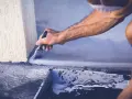 Mężczyzna maluje pędzlem szarą farbą betonową przy ścianie. Widać kuwetę z farbą.