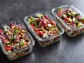 Trzy szklane pojemniki wypełnione kolorową sałatką z ciecierzycą, ogórkiem, pomidorem, czerwoną cebulą i fetą. Idealna na lunch.