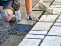 Ręce w rękawiczkach układają kamienne kostki na podsypce. Widać młotek i sznur geodezyjny. Prawidłowe suchy beton proporcje to klucz do trwałej nawierzchni.