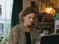 Kvinna med lockigt hår sitter vid en laptop och ska skriva mail. Hon bär en blommig skjorta och ett guldfärgat halsband.