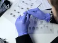 Ein Forensiker untersucht eine Patrone mit einer Pinzette. Im Hintergrund sind Fingerabdrücke zu sehen.