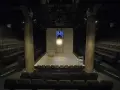 Pusta scena w **teatrze stu** miejsc, z rzędami siedzeń po bokach. Na scenie łóżko, białe krzesło i reflektory.