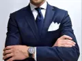 Elegancki mężczyzna w granatowym garniturze, białej koszuli, z granatowym krawatem w kropki i białą poszetką. Zegarek na ręce. Idealny przykład, jaki krawat do granatowego garnituru.