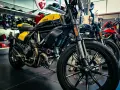 Ducati Scrambler gialla e nera, pronta per i raduni moto. Dietro, altre moto e abbigliamento tecnico.