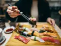Eine Hand hält mit Stäbchen ein Stück Sushi mit gehacktem Thunfisch. Auf dem Teller liegen verschiedene Sushi-Rollen und Nigiri, bereit für den Genuss in Berlin.