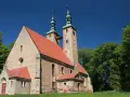 Stary kościół w stolcu z dwiema wieżami i zielonymi dachami, otoczony bujną zielenią drzew pod błękitnym niebem.