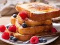 Una pila de tostadas francesas doradas, decoradas con frambuesas y arándanos, perfectas para aprender cómo se hacen las tostadas francesas.
