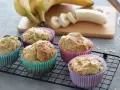 Fünf leckere Bananen Muffins in bunten Förmchen, bereit für Kinder. Bananen und Mehl sind im Hintergrund zu sehen.