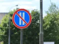 Znak B38 zakazujący postoju. Okrągły znak z czerwonym obramowaniem, niebieskim tłem i dwoma białymi pionowymi pasami przekreślonymi czerwoną linią.