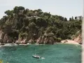 A sailboat floats in turquoise water near a rocky, tree-covered cliff with buildings. Perfect for weekend trips from Barcelona by train.