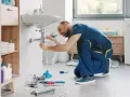 Instalator klęczy przy umywalce, pokazując, jak wymienić kran. Narzędzia i części są rozłożone na podłodze.