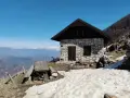 Bivacco Monte Grappa, rifugio in pietra con tavolo da picnic e vista sulle montagne innevate.