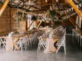 Drewniana stodoła udekorowana na przyjęcie. Długie stoły z kwiatami i świecami, wiszące dekoracje z traw. Widać różnicę między stylem boho chic a rustykalnym w dekoracjach kwiatowych.