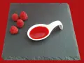 Quatre framboises fraîches à côté d'une cuillère blanche remplie de coulis de framboise, sur une ardoise.