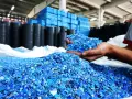 Ręce trzymające niebieski granulat plastiku, który jest efektem polimeryzacji. W tle stosy czarnych i niebieskich pojemników.