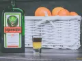 Butelka Jägermeistera i shot z dwoma warstwami, obok koszyk z pomarańczami. Idealne na wieczór, jak pić jagermeister.
