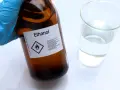 Braune Glasflasche mit der Aufschrift "Ethanol" und einem Warnsymbol. Daneben ein Becherglas mit klarer Flüssigkeit. Hier gibt es 100 prozentigen Alkohol.