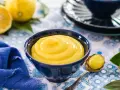 Una ciotola blu piena di vellutata crema al limone, con limoni freschi e un tovagliolo blu sullo sfondo.