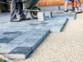 Układanie nowoczesnych płyt betonowych, co zamiast kostki brukowej. Pracownicy z taczką tworzą ścieżkę.