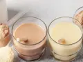 Dwa szklanki z napojami proteinowymi, obok proszek białkowy w miarkach i miseczkach.