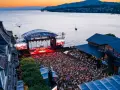 Ein großes Publikum feiert bei Sonnenuntergang ein Konzert am See. Die Bühne des Jazz Montreux Festivals ist beleuchtet, im Hintergrund der Genfersee.