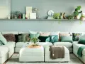 Sofá modular claro con cojines verdes y estampados, y una estantería con plantas y objetos decorativos, incluyendo una casita de cristal con **muebles pintados en verde**.