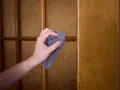Mano limpiando con paño gris una puerta de madera con paneles de vidrio texturizado. Guía práctica sobre como limpiar las puertas de madera.