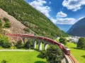 Czerwony pociąg Bernina Express przejeżdża przez kamienny wiadukt w Alpach. Sprawdź bernina express cena, by zaplanować podróż.