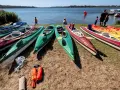 Kajaki czekają nad jeziorem. Kilka osób przygotowuje się do wodnych przygód, a w tle widać łodzie i żaglówki.