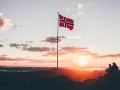 Zachód słońca w Norwegii. Dwie osoby siedzą na skale, patrząc na krajobraz. Flaga Norwegii powiewa na maszcie. Czy do Norwegii można lecieć na dowód osobisty?