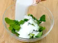 Zucchero bianco viene versato su foglie di menta fresca in una ciotola di vetro, per preparare uno sciroppo di menta fatto in casa.