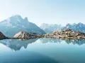 Un lac alpin reflète des montagnes majestueuses, un aperçu des plus belles randonnées de France. Une personne se tient sur la rive rocheuse.