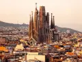 Vue panoramique de Barcelone au coucher du soleil, avec la Sagrada Familia au centre. Les travaux de la Sagrada Familia continuent, sa silhouette imposante se détachant sur le ciel orangé.