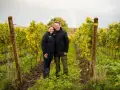 Ein Paar steht lächelnd in einem Weinberg in Schleswig-Holstein. Im Hintergrund ist ein Regenbogen zu sehen.