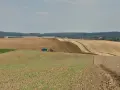 Czerwony traktor z przyczepą nawozi pole, przygotowując je do dalszej rekultywacji. W tle widać kolejne pola i las.