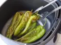 Pimientos verdes asándose en una freidora de aire. Unas pinzas metálicas sujetan uno de los pimientos, mostrando su piel ligeramente chamuscada.