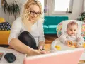 Uśmiechnięta mama pracuje przy laptopie, podczas gdy dziecko bawi się w krzesełku. Planuje zwrot podatku na dziecko.